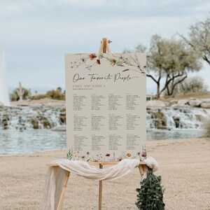 以下が含まれることがあります： 花柄の縁取りが施されたベージュの結婚式席次表。表には「Our Favorite People」というタイトルと、エミリーとジョシュアの2人の名前、2023年9月24日の日付が記載されています。表には、ゲストの名前と割り当てられたテーブルが記載されています。