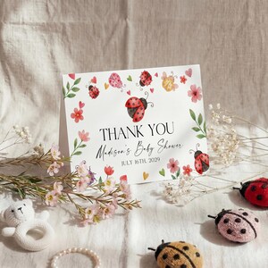 May include: A baby shower thank you card with ladybug and floral illustrations. The card reads "THANK YOU Madison's Baby Shower JULY 16th, 2029". Decorative ladybug plush toys and flowers are arranged around the card.