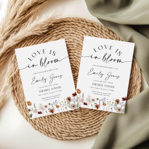 May include: Two white wedding invitations with floral accents and the text "Love is in bloom." The invitations include the name "Emily Jones," event details, and venue information. The cards are on a woven mat.