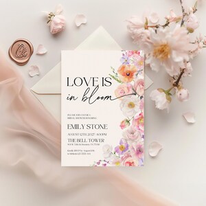May include: A white card with a floral design border. The text "LOVE IS in bloom" is in black script font. The card is an invitation to a bridal shower for Emily Stone on August 12th, 2027 at 4:00 PM at The Bell Tower.