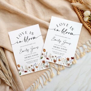 May include: Two white wedding invitations with the text "Love is in bloom" and the name "Emily Jones" in elegant script. Floral illustrations in shades of brown, red, and yellow adorn the bottom. The invitations are on a beige fabric.