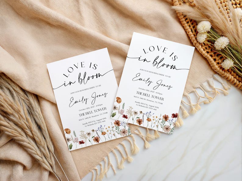 May include: Two white wedding invitations with the text "Love is in bloom" and the name "Emily Jones" in elegant script. Floral illustrations in shades of brown, red, and yellow adorn the bottom. The invitations are on a beige fabric.