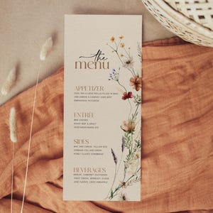 Puede incluir: Una tarjeta de menú beige con un diseño floral y las palabras "the menu" en escritura cursiva marrón. El menú enumera aperitivos, platos principales, acompañamientos y bebidas.
