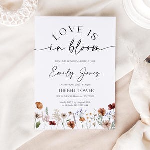 May include: A white invitation card with the text "LOVE IS in bloom" in elegant script. The card includes details for a wedding, with the bride's name, date, time, and location, and a floral border at the bottom.