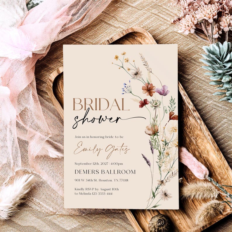 A minimalist bridal shower invitation template with a light beige background. On the right side, there is a delicate watercolor arrangement of wildflowers and dried grasses in earthy tones of rust, purple, and cream. The typography is a mix of modern serif and elegant script fonts.