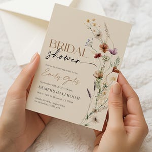 A minimalist bridal shower invitation template with a light beige background. On the right side, there is a delicate watercolor arrangement of wildflowers and dried grasses in earthy tones of rust, purple, and cream. The typography is a mix of modern serif and elegant script fonts.