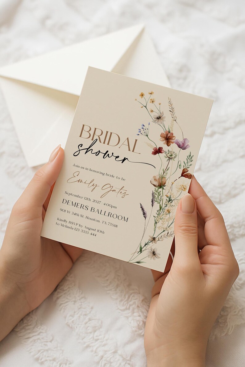 A minimalist bridal shower invitation template with a light beige background. On the right side, there is a delicate watercolor arrangement of wildflowers and dried grasses in earthy tones of rust, purple, and cream. The typography is a mix of modern serif and elegant script fonts.