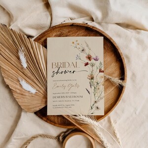 A minimalist bridal shower invitation template with a light beige background. On the right side, there is a delicate watercolor arrangement of wildflowers and dried grasses in earthy tones of rust, purple, and cream. The typography is a mix of modern serif and elegant script fonts.