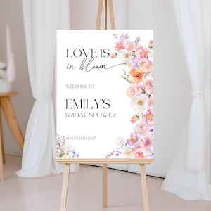 Op de afbeelding: Een wit bord met de tekst "Love is in bloom" en een bloemige rand. Het bord vermeldt ook "Welkom bij Emily's Bridal Shower" en "12 augustus 2027".