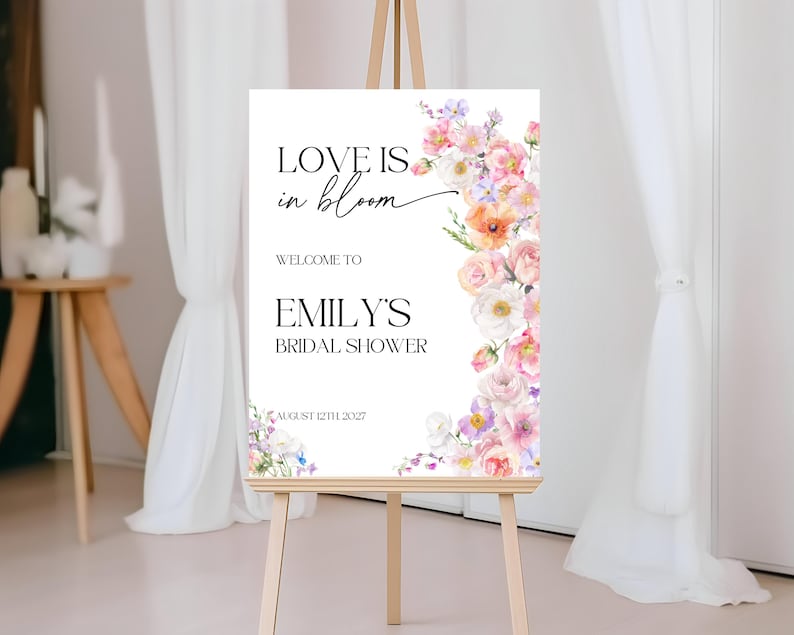Op de afbeelding: Een wit bord met de tekst "Love is in bloom" en een bloemige rand. Het bord vermeldt ook "Welkom bij Emily's Bridal Shower" en "12 augustus 2027".