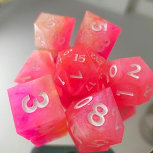 May include: A set of seven polyhedral dice in pink and orange hues. The dice feature white numbering and are made from a translucent material. The dice are arranged in a pile, with some partially hidden by others.