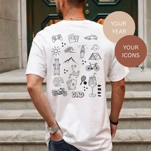 Peut inclure: Un t-shirt blanc avec un motif graphique en lignes noires au dos. Le design comprend diverses icônes telles qu'une manette de jeu, une voiture, une planche de surf, une tente, un vélo, une moto, une part de pizza, des cartes et le mot DAD avec 1989.