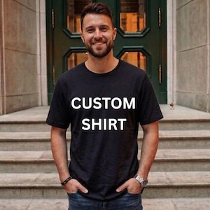 Könnte beinhalten: Schwarzes T-Shirt mit den Worten "CUSTOM SHIRT" in weißen Blockbuchstaben. Das T-Shirt wird mit blauen Jeans getragen. Die Person steht vor einer grünen Tür und Steinstufen.