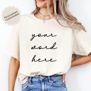 Può includere: T-shirt color crema con le parole "your word here" in un carattere nero. La maglietta è abbinata a jeans blu. Una piccola etichetta colorata nell'angolo in alto a sinistra recita "Personalisert Customizable".
