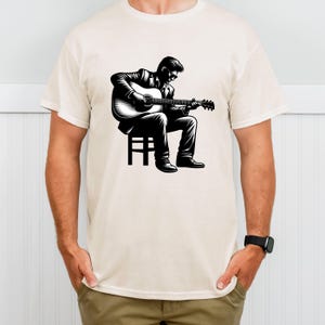 May include: Cream-colored t-shirt featuring a black and white illustration of a person playing an acoustic guitar while seated on a stool. The illustration depicts the person wearing a suit and sunglasses.
