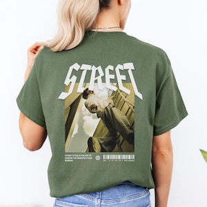 Könnte beinhalten: Olivgrünes T-Shirt mit Grafik auf dem Rücken. Die Grafik zeigt das Wort "STREET" in einer kräftigen, stilisierten Schriftart über einem Bild einer Person. Unter dem Bild steht der Text "STREET STYLE IS THE ART OF MAKING THE SIDEWALK YOUR RUNWAY."