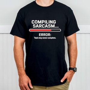 Könnte beinhalten: Schwarzes T-Shirt mit weißem Text "COMPILING SARCASM..." über einem roten und schwarzen Fortschrittsbalken. Darunter steht "ERROR: Task may never complete."