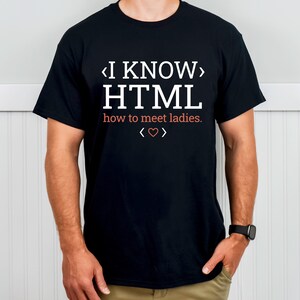 Op de afbeelding: Zwart T-shirt met witte tekst: "I KNOW HTML" boven "how to meet ladies." Een hartsymbool staat onder de tekst. Het shirt heeft korte mouwen en lijkt van katoen te zijn.
