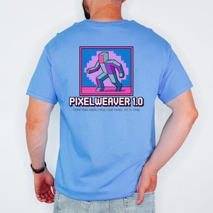 Könnte beinhalten: Hellblaues T-Shirt mit einem pixeligen Figuren-Design in einem rosa und blauen Quadrat. Unter der Grafik steht der Text "PIXELWEAVER 1.0" mit dem Slogan "CRAFTING REALITIES, ONE PIXEL AT A TIME."