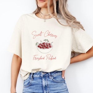 Könnte beinhalten: Cremefarbenes T-Shirt mit roter Grafik einer Schale mit Kirschen und dem Text "Sweet Cherries, Freshly Picked". Das Design ist im handschriftlichen Stil gehalten. Das Shirt besteht aus einem weichen Material.
