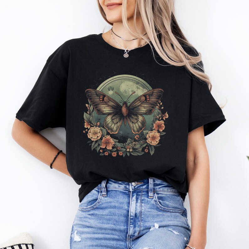 May include: Black t-shirt with a butterfly graphic. The butterfly is brown and tan, set against a green moon and surrounded by flowers. The shirt is a casual style, suitable for everyday wear.
