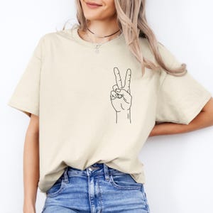 Könnte beinhalten: Ein cremefarbenes T-Shirt mit einer schwarzen Strichzeichnung einer Hand, die das Friedenszeichen zeigt. Die Grafik befindet sich auf der linken Brustseite. Das Shirt wird mit einer blauen Jeans kombiniert.