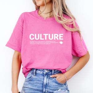 Pode incluir: Uma t-shirt rosa brilhante com a palavra "CULTURE" em letras brancas grandes. Abaixo, uma descrição da cultura e do século XX. A t-shirt é combinada com jeans azuis.