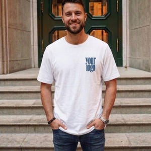 Könnte beinhalten: Ein weißes kurzärmeliges T-Shirt mit dem Text "your logo" in blauer Serifenschrift auf der linken Brustseite. Das T-Shirt hat einen Rundhalsausschnitt und wird mit einer dunkelblauen Jeans, einer silbernen Armbanduhr und einem schwarzen Perlenarmband getragen.