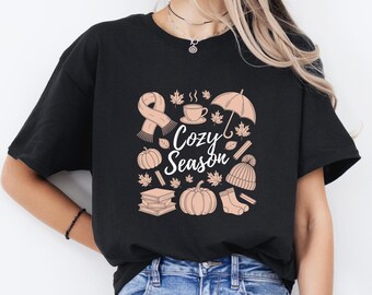 Könnte beinhalten: Schwarzes T-Shirt mit einem "Cozy Season"-Motiv in weißer Schrift. Das Design umfasst einen Schal, eine Kaffeetasse, einen Regenschirm, Ahornblätter, Kürbisse, Bücher, eine Mütze und Socken in einem weichen Pfirsichton.