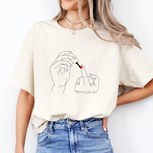 Puede incluir: Una camiseta de manga corta color crema con un gráfico de líneas negras de dos manos. Una mano sostiene un frasco de esmalte de uñas, mientras que la otra mano está extendida con un dedo levantado, mostrando un pequeño punto rojo de esmalte.