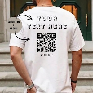 Könnte beinhalten: Ein weißes T-Shirt mit dem schwarzen Text YOUR TEXT HERE auf der Rückseite, platziert über einem großen schwarz-weißen QR-Code. Unter dem QR-Code steht der Text SCAN ME! in Schwarz. Eine kreisförmige Grafik zeigt Optionen für individuellen Text.