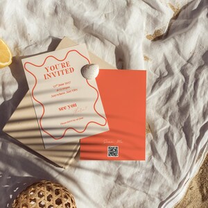 Puede incluir: Un conjunto de invitaciones con una tarjeta beige con texto rojo que dice "YOU'RE INVITED" y una tarjeta naranja. La invitación está decorada con una concha. La escena incluye rodajas de limón, una bola tejida y una tela blanca sobre arena.