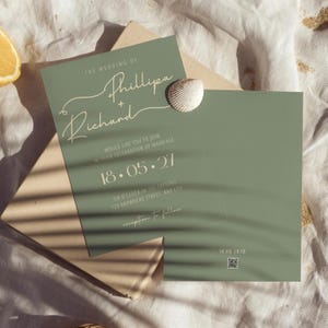 May include: Sage green wedding invitation with elegant script text. The invitation includes the names "Phillipa + Richard", the date "18.05.27", and event details. A seashell rests on the invitation, which is on top of a neutral envelope.
