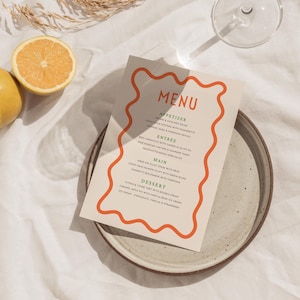 May include: A restaurant menu with an orange wavy border, listing appetizers, entrées, main courses, and desserts. The menu is on a ceramic plate, with a lemon and a half-cut orange nearby.