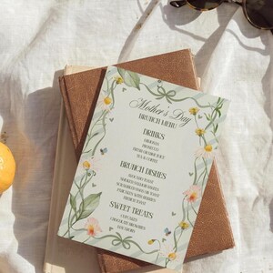 Peut inclure: Un menu de brunch pour la fête des mères avec des accents floraux et des rubans. Le menu répertorie les boissons, les plats du brunch et les douceurs. Le menu est sur deux livres et un tissu blanc.