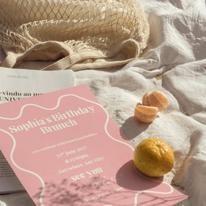 May include: A pink invitation with the text "Sophia's Birthday Brunch" and event details, alongside a whole lemon and orange segments. A beige net bag and a book are also present, all arranged on a white cloth.