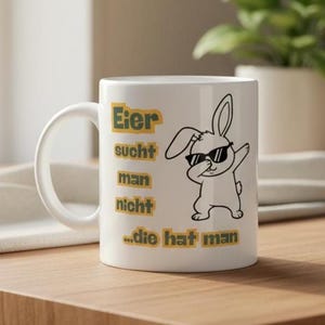 Könnte beinhalten: Weiße Keramik-Tasse mit Henkel, mit dem Text "Eier sucht man nicht ...die hat man" in Gelb und einem schwarzen Cartoon-Hasen mit Sonnenbrille. Die Tasse steht auf einer Holzoberfläche.