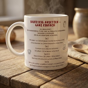 Könnte beinhalten: Eine weiße Keramiktasse mit deutschem Text und Anweisungen zur Herstellung von Sauerteig. Die Tasse steht auf einer Holzoberfläche, aus der Dampf aufsteigt. Ein Holzlöffel und ein Keramiktopf befinden sich im Hintergrund.