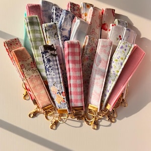 May include: A collection of fabric wrist lanyards in various patterns and colors, including floral, gingham, and solid designs. Each lanyard has a gold-colored clasp for attaching keys or other items.