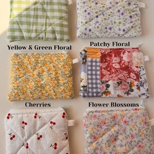 May include: Six small, quilted pouches with zippers, each featuring a unique fabric design. Patterns include green gingham, purple flowers, yellow and green floral, patchwork floral, cherries, and flower blossoms. The pouches are arranged on a white surface.