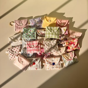 May include: A collection of small fabric pouches with various floral and patterned designs. The pouches have a button closure and a gold ring attached.