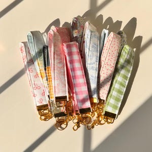 May include: A collection of fabric wrist keychains in various patterns and colors. The keychains feature floral, gingham, and dotted designs in shades of pink, blue, and green. Each keychain has a gold-colored clasp and hardware.