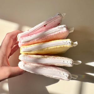 May include: A stack of four small zippered pouches in various pastel colors and patterns. The pouches are made of soft fabric and have white zippers.