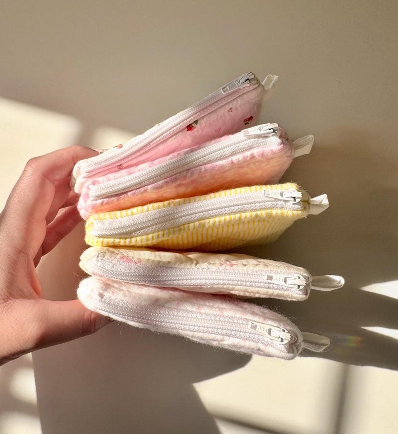 May include: A stack of four small zippered pouches in various pastel colors and patterns. The pouches are made of soft fabric and have white zippers.
