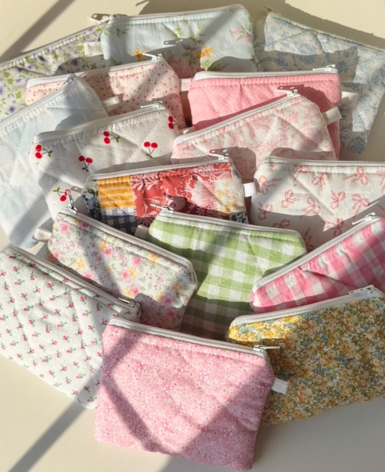 May include: A collection of small, quilted fabric pouches in various floral, cherry, and gingham patterns. The pouches are in shades of pink, white, green, and yellow, with zippers and a quilted texture. The pouches are ideal for storing small items.