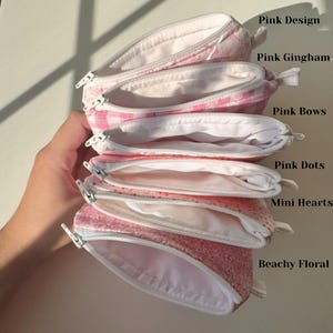 May include: A stack of open, pink zippered pouches with white interiors. The pouches feature various patterns, including pink gingham, dots, mini hearts, and floral designs. The text on the image reads "Pink Design", "Pink Gingham", "Pink Bows", "Pink Dots", "Mini Hearts", and "Beachy Floral".