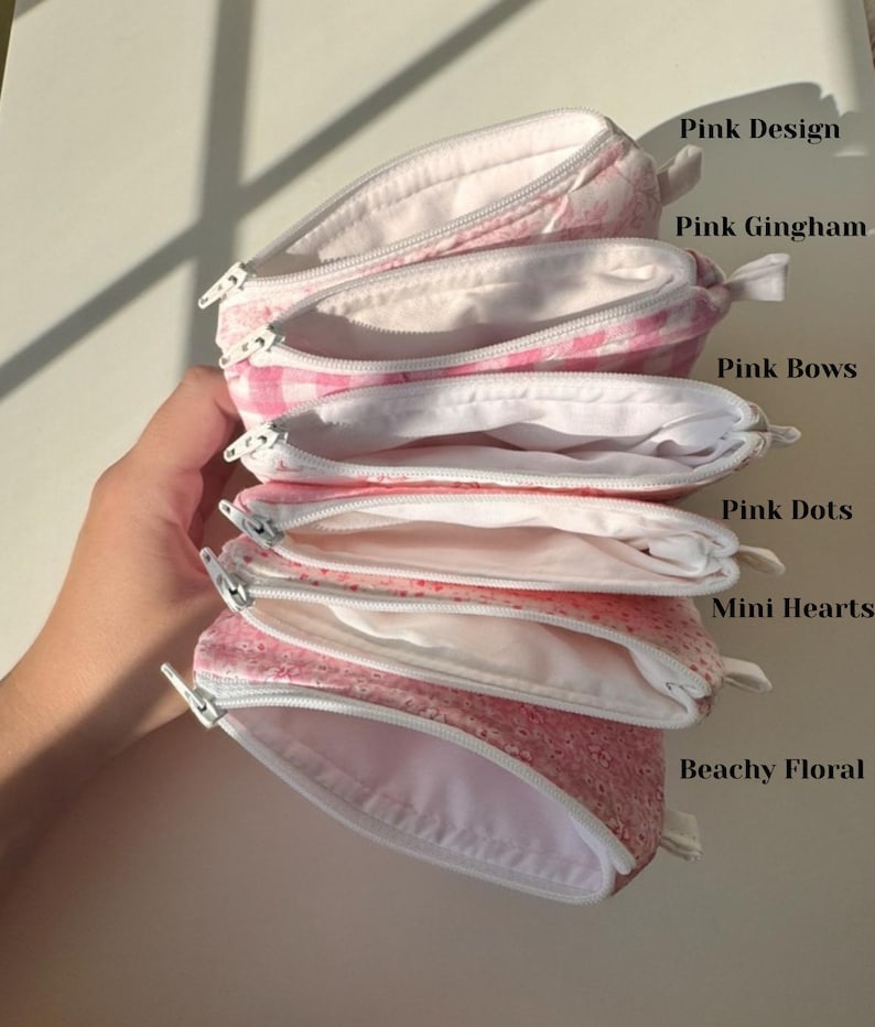 May include: A stack of open, pink zippered pouches with white interiors. The pouches feature various patterns, including pink gingham, dots, mini hearts, and floral designs. The text on the image reads "Pink Design", "Pink Gingham", "Pink Bows", "Pink Dots", "Mini Hearts", and "Beachy Floral".