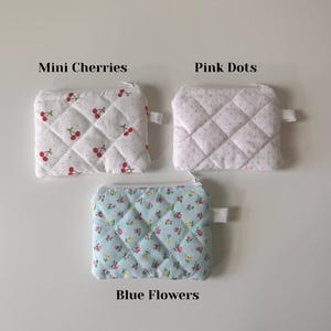 May include: Three quilted zipper pouches with different patterns. The first pouch is white with red cherries, the second is white with pink dots, and the third is light blue with small pink and yellow flowers. The pouches are labeled "Mini Cherries", "Pink Dots", and "Blue Flowers".