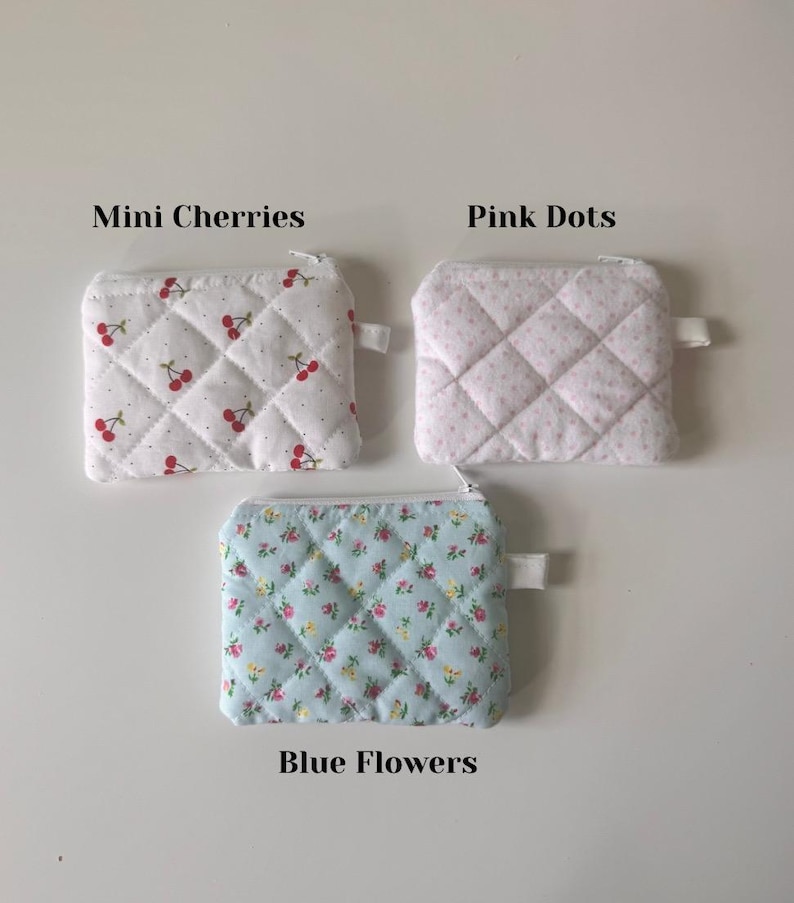 May include: Three quilted zipper pouches with different patterns. The first pouch is white with red cherries, the second is white with pink dots, and the third is light blue with small pink and yellow flowers. The pouches are labeled "Mini Cherries", "Pink Dots", and "Blue Flowers".