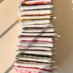 May include: A stack of small, rectangular fabric pouches with white zippers. The pouches feature various floral and patterned designs in shades of pink, red, yellow, blue, and green. The pouches are stacked vertically against a neutral background.
